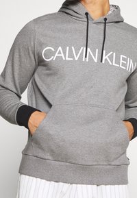 Sweatshirt gris avec une grande impression blanche "CALVIN KLEIN" sur la poitrine, poignets côtelés noirs et poche kangourou, featuring une capuche doublée.