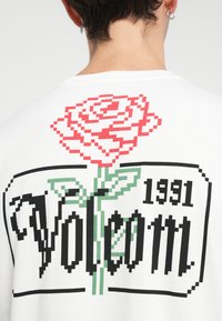 T-shirt blanc avec un graphique de rose rouge pixellisé et le texte noir "Volcom 1991" avec des feuilles vertes, sur le dos. En matériau coton.
