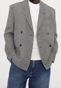 Mężczyzna w szaro-skalowanym, w kratkę dwurzędowym marynarkę na białej koszuli, z niebieskimi dżinsami, z rękami w kieszeniach, na jednolitym tle.