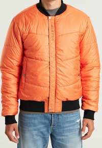 Man in een feloranje gewatteerde jas met zwarte manchetten en tailleband, gecombineerd met lichtblauwe versleten jeans.