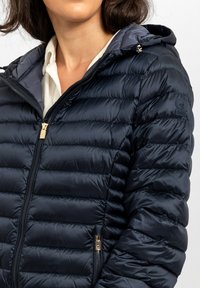 Giacca imbottita blu navy con chiusura frontale a zip, texture a coste e cappuccio. Presenta dettagli in metallo dorato e un logo sulla manica sinistra.