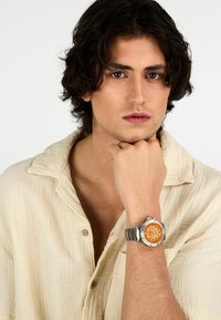 Orologio in metallo argento con quadrante arancione, dotato di indicatori delle ore neri e bracciale in acciaio inossidabile, indossato da una persona con una camicia beige chiara.