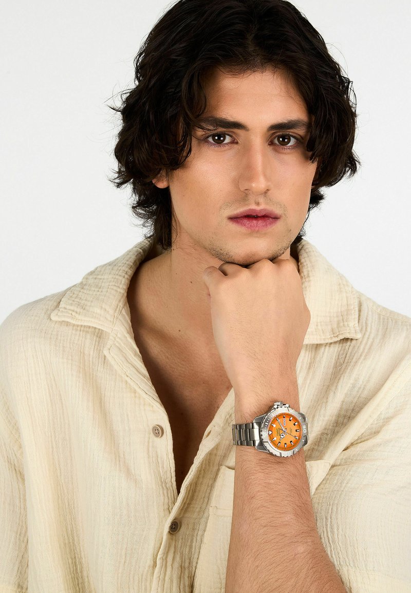 Orologio in metallo argento con quadrante arancione, dotato di indicatori delle ore neri e bracciale in acciaio inossidabile, indossato da una persona con una camicia beige chiara.