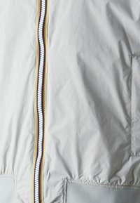 Giacca grigia chiara con zip frontale e dettagli colorati, orlo a costine. Include una tasca laterale con una texture liscia.