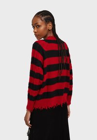 Červený a čierny pruhovaný sveter s voľným strihom, rozstrapkaným lemom a textúrovaným úpletom, nosený osobou s vrkôčikmi, otočenou dozadu.