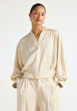 Donna che indossa una camicetta in raso color crema, a taglio ampio, e pantaloni abbinati con piccoli pois neri, in piedi con le mani nelle tasche.