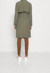 Trencz w oliwkowej zieleni z paskiem w talii, długimi rękawami i klapą z tyłu, zestawiony z brązowymi spodniami i białymi butami na kwadratowym obcasie.