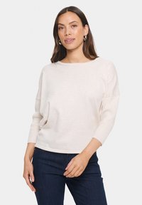 Maglione beige chiaro con una texture morbida, scollo rotondo e maniche a tre quarti, abbinato a jeans in denim scuro.