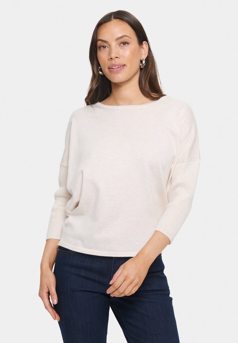 Maglione beige chiaro con una texture morbida, scollo rotondo e maniche a tre quarti, abbinato a jeans in denim scuro.