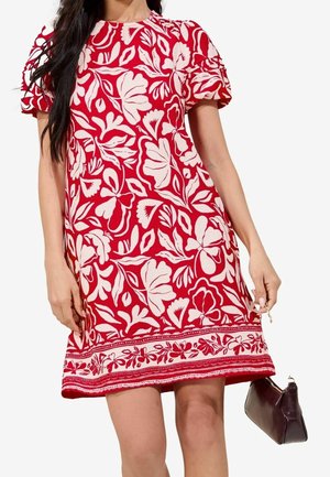 Kobieta w czerwonej sukience z krótkim rękawem w białe kwiatowe wzory, trzymająca małą ciemnobrązową torebkę na białym tle.
