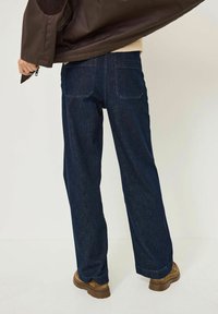 Tmavě modré denimové široké kalhoty se dvěma zadními kapsami, v kombinaci s hnědou svrchní bundou. Látka má hladkou texturu a strukturovaný střih.