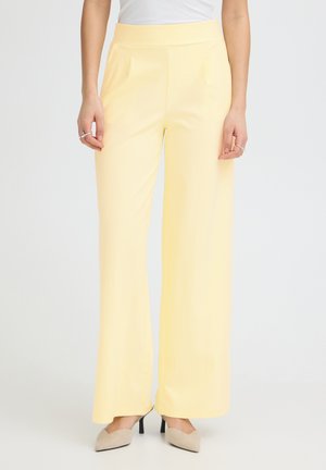 Donna che indossa pantaloni giallo pallido a vita alta e gamba larga, top bianco e décolleté beige con punta a mandorla, in piedi davanti a uno sfondo semplice.