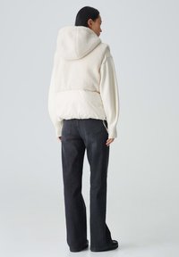 Cremfarbene Kapuzenjacke mit einem strukturierten Fleece-Oberteil und glattem Unterteil, kombiniert mit hochgeschnittenen, dunkelblauen Jeans und schwarzen Schuhen.