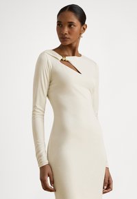 Vestido creme de manga comprida com um design ajustado, apresentando um corte diagonal adornado com um detalhe dourado perto do decote. Textura suave.