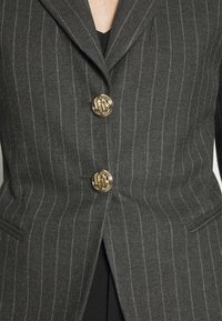 Blazer gris foncé à fines rayures avec deux boutons décoratifs dorés fermés, laissant apparaître partiellement un vêtement noir en dessous.