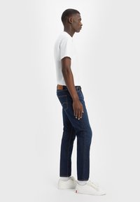 Biała koszulka z krótkim rękawem zestawiona z ciemnoniebieskimi dżinsami o prostym kroju, noszona z białymi trampkami. Dżinsy mają brązową skórzaną metkę.