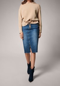 Kvinna i en beige huvtröja, knälång blå denimkjol med framklippning och svarta mockastövletter, står framför en grå bakgrund.