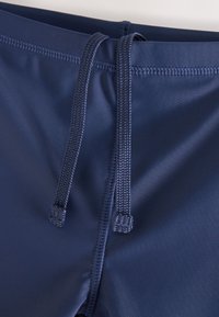 Nahaufnahme von marineblauen Badehosen mit geflochtenem Kordelzug und Flatlock-Nähten rund um den Bund und den Schrittbereich.