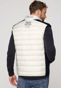 Gilet imbottito senza maniche con parte anteriore di un leggero crema, retro blu scuro e polsini a coste; presenta il logo sul colletto e sul retro.