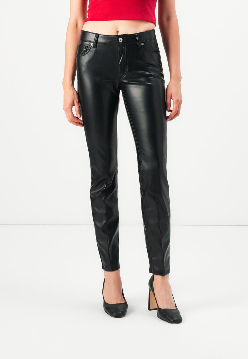 Vrouw in zwarte leren slim-fit broek, zwarte hoge hakken en een rode crop top tegen een witte achtergrond.