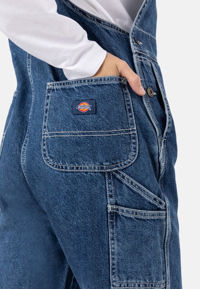 Salopettes en denim bleu foncé avec des coutures claires, munies d'une poche avant et d'un patch de marque. Le tissu semble résistant avec une texture lisse.