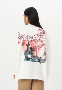 Sweatshirt blanc surdimensionné avec un design aquarelle coloré de fleurs roses, d'une maison et de poissons koi au dos. Matière douce en coton.