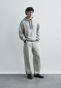 Ανοιχτό γκρι φούτερ με κουκούλα και υφασμάτινη σχεδίαση, σετ με φαρδιά ανοιχτά μπλε τζιν και μαύρα δερμάτινα παπούτσια, στέκεται μπροστά σε ουδέτερο φόντο.