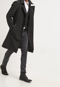 Schwarzer Übermantel mit aufgeschlagenem Kragen, einreihigem Design und seitlichen Taschen, getragen über einer schwarzen Weste und grauen Hosen, kombiniert mit schwarzen Stiefeln.
