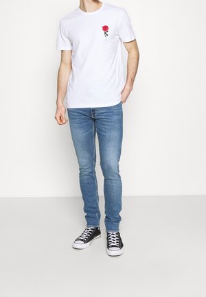 Camiseta blanca de algodón con un bordado de rosa roja en el pecho izquierdo, combinada con jeans ajustados de mezclilla azul claro y zapatillas negras.