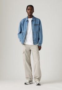 Jeansová bunda vo svetlom modrom odtieni, s elastickými manžetami a zipsovým vreckom na hrudi. Skombinovaná s béžovými cargo nohavicami a viacfarebnými teniskami.