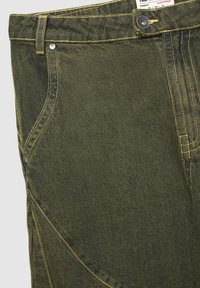 Pantaloni in denim verde scuro con cuciture gialle a contrasto, le caratteristiche includono una chiusura a bottone, passanti per cintura e tasche frontali.