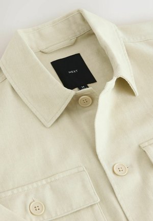 Beige Hemd zum Zumachen mit spitzem Kragen, Brusttasche und schwarzem Markenlabel mit der Aufschrift "NEXT" im Nackenbereich.