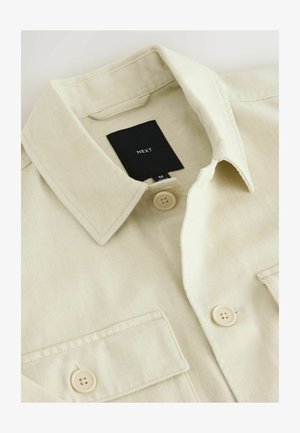Beige Hemd zum Zumachen mit spitzem Kragen, Brusttasche und schwarzem Markenlabel mit der Aufschrift "NEXT" im Nackenbereich.