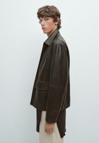 Brown leather jacket with a collared neckline, two front pockets, and a relaxed fit, worn over a textured cream garment.