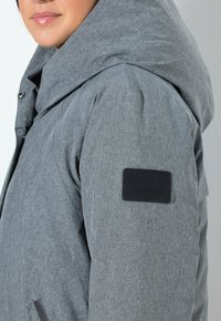 Chaqueta acolchada gris con un acabado texturizado, que cuenta con una gran capucha, bolsillos laterales con cremallera y una etiqueta de marca negra en la manga izquierda.