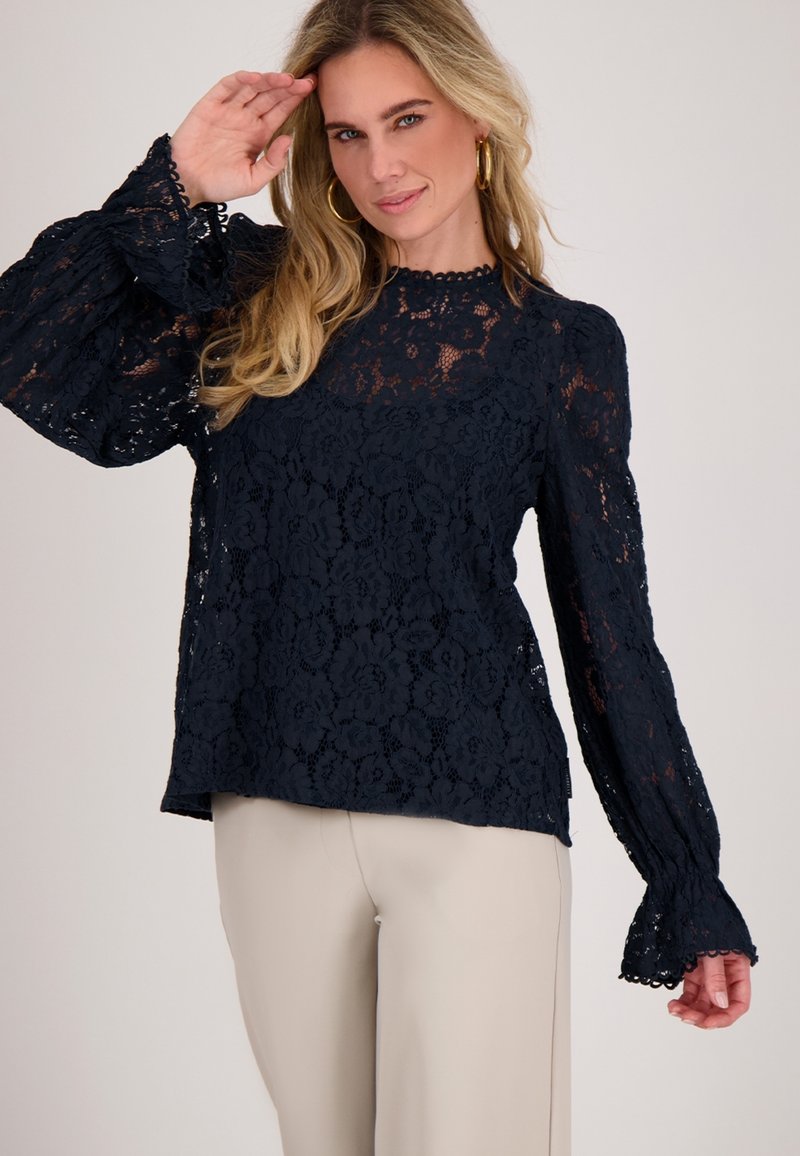 Marineblauwe kanten blouse met lange, volumineuze mouwen met gegolfde randen, bloemmotieven en een ronde halslijn. Gecombineerd met beige broek.