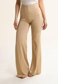 Pantaloni beige a vita alta realizzati in tessuto liscio con un design a gamba svasata e cuciture sottili, dotati di chiusura con zip laterale.