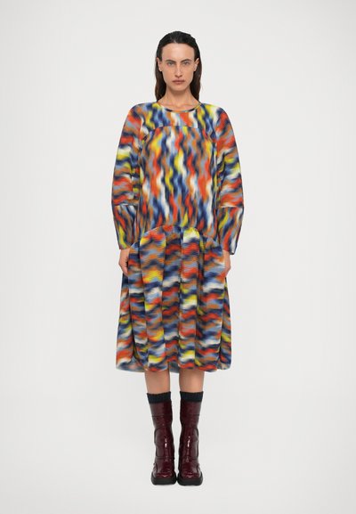 Femme portant une robe à manches longues à motif ondulé multicolore avec une jupe plissée, associée à des bottes bordeau mi-mollet et des chaussettes noires.