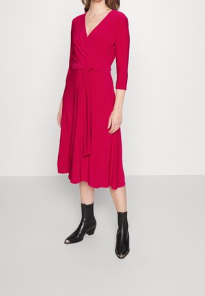 Femme portant une robe portefeuille fuchsia à manches longues et longueur genou, avec une ceinture nouée à la taille, assortie à des bottines noires à bouts pointus.