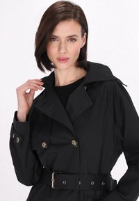 Czarny trenchcoat z paskiem w talii, podwójnymi guzikami, ostrym kołnierzem i miękką teksturą materiału. Posiada subtelne akcenty z elementów metalowych.