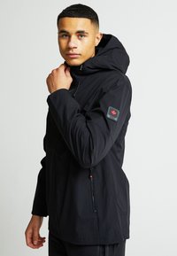 Černá voděodolná bunda s kapucí, zipovými kapsami a nášivkou na rukávu. Hladký povrch a nastavitelné velikosti.