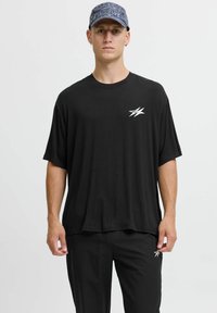 Camiseta negra de gran tamaño con mangas cortas y escote redondo, que presenta un logo blanco en el lado izquierdo del pecho; combinada con pantalones negros.