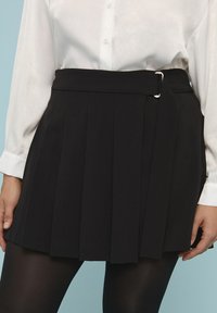Falda corta plisada negra con una textura suave, que presenta una hebilla lateral y una cintura ajustada. Se lleva con una camisa blanca de botones y medias.