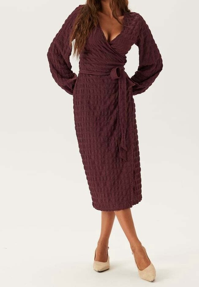 Femme portant une robe portefeuille texturée d'un violet profond avec des manches longues et une taille nouée, associée à des talons beiges pointus.