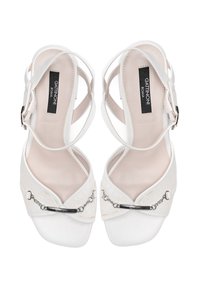 White heeled sandals made of leather, featuring a crisscross strap design, metallic chain accent, and smooth texture on the sole.
