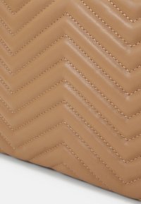 Torebka w kolorze tan, z chevronowym pikowaniem, widocznymi szwami, gładką strukturą i wyraźnym kształtem.