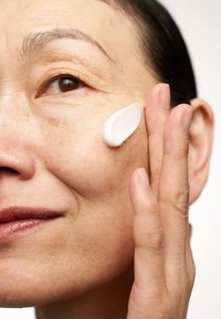 Een persoon die een witte creme op zijn wang aanbrengt, met gladde huid en fijne lijntjes, tegen een neutrale achtergrond die het product benadrukt.