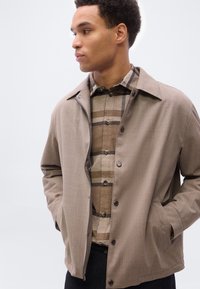 Άνδρας με μπεζ σακάκι πάνω από καφέ καρό πουκάμισο, τα χέρια στις τσέπες του σακακιού, κοιτάζει προς τα αριστερά του με φόντο απλό ανοιχτόχρωμο.