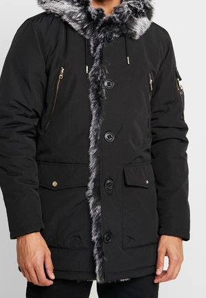 Manteau d'hiver noir avec bordure en fausse fourrure grise sur la capuche et le devant, fermeture par boutons et zip, et plusieurs poches à l'avant et sur la manche.