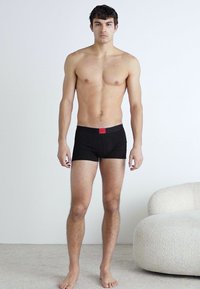 Schwarze Baumwoll-Boxershorts mit einem roten Logo-Bund. Das Modell steht auf einem hellen Teppich und präsentiert ein eng anliegendes Design.